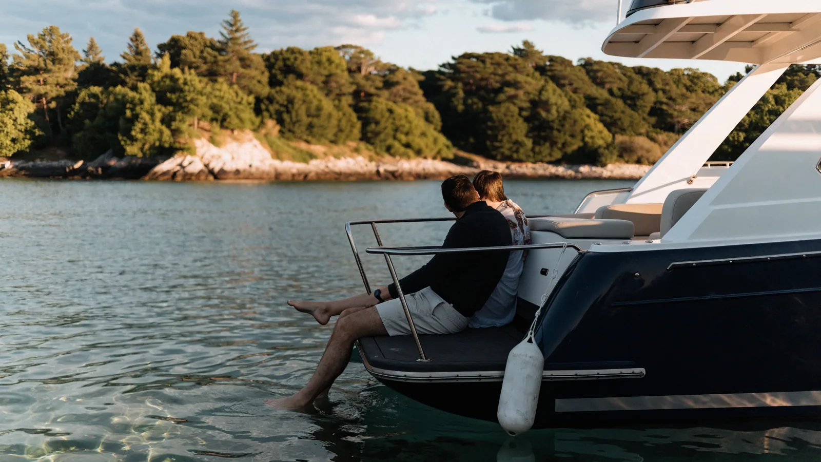 Una pareja sentada al atardecer en la plataforma de baño de su yate a motor, en una cala tranquila, con los pies en el agua