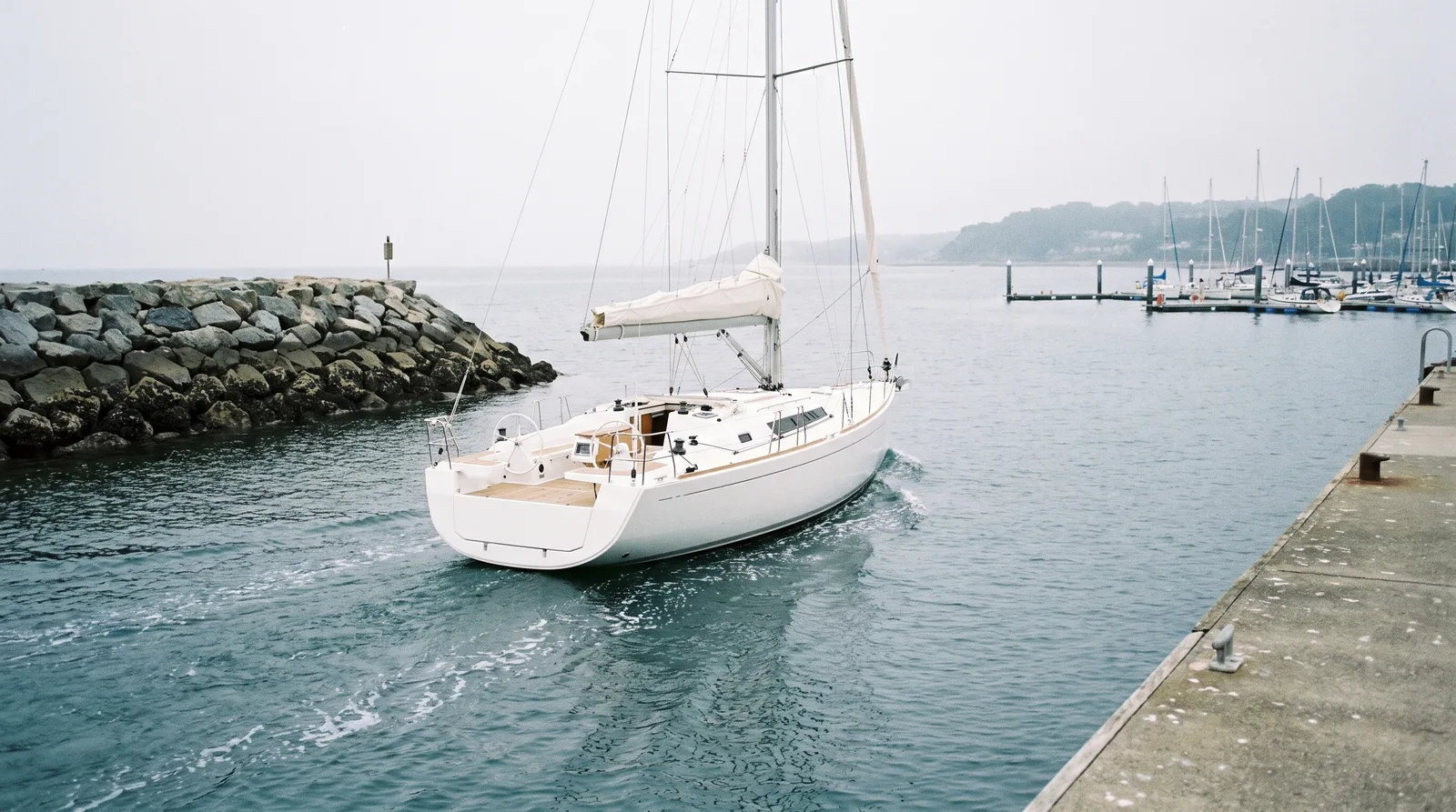 Un velero blanco con las velas recogidas sale a motor del puerto junto a un espigón de roca, con un muelle de hormigón y pantalanes entre la bruma al fondo.