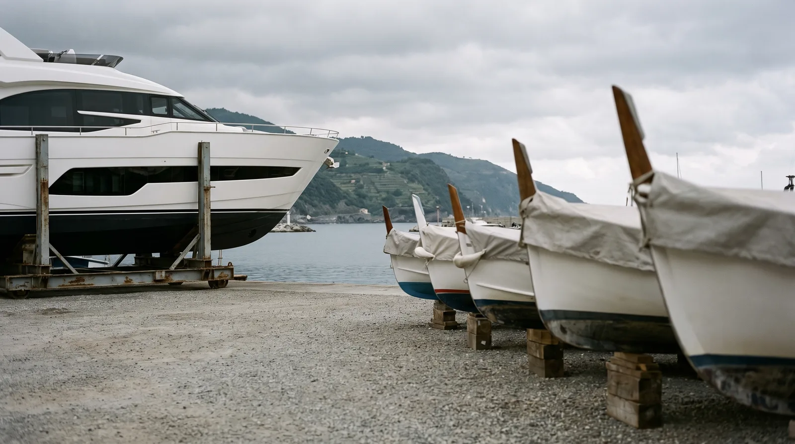 Explanada de un astillero en la costa de Liguria: a la izquierda un yate a motor blanco y nuevo sobre una cuna de transporte de acero, a la derecha una fila de pequeñas barcas sobre tacos de madera con lonas grises, al fondo el agua del puerto y colinas en terrazas.