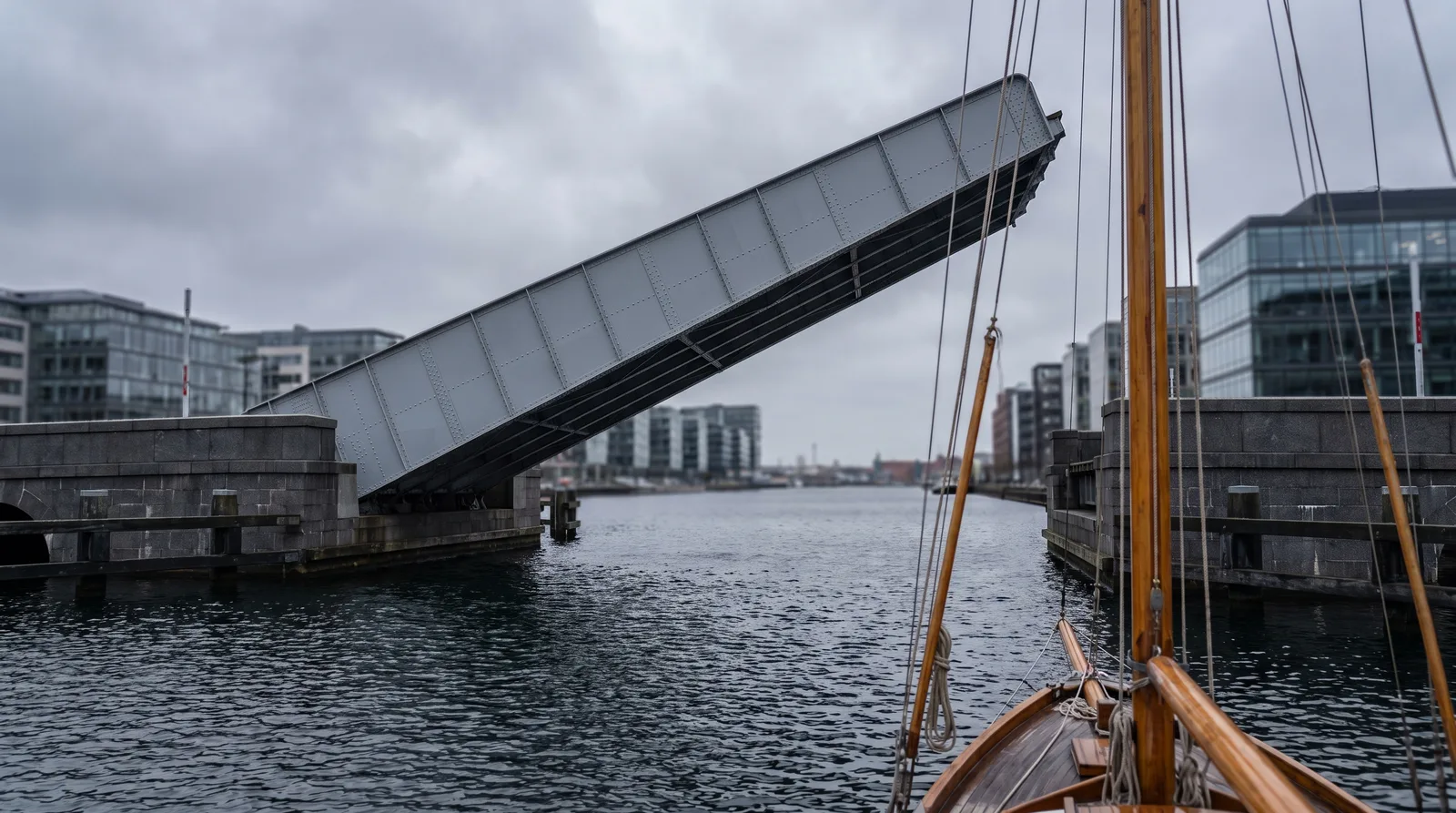 La hoja de acero levantada de un puente basculante urbano sobre agua oscura, con el mastil de madera de un velero pequeno desenfocado delante.
