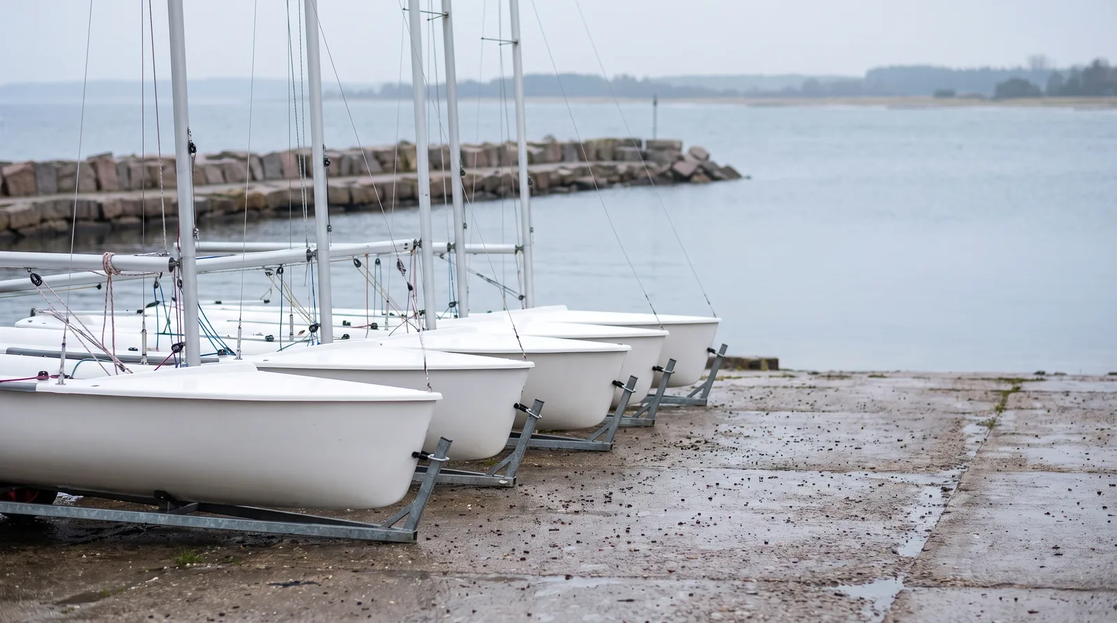 Una hilera de botes de regata blancos sobre carros de varada galvanizados en una rampa de hormigón mojada, con los mástiles arbolados y sin velas, y detrás un espigón de piedra y una bahía báltica tranquila y gris con una costa baja y arbolada entre la bruma