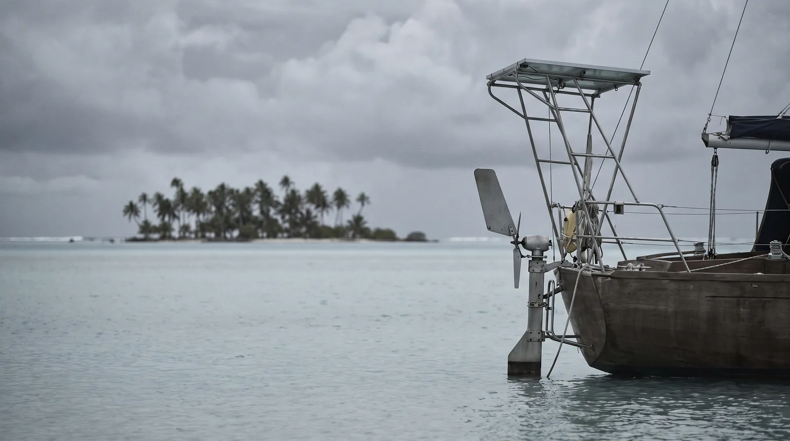 La popa de un velero de crucero fondeado en una laguna amplia, con un piloto de viento en el espejo y una placa solar sobre un arco de acero inoxidable. Al fondo, un islote de cocoteros bajo un cielo cubierto.