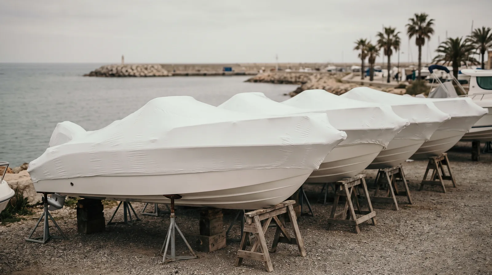 Una fila de lanchas nuevas envueltas en film blanco sobre caballetes de madera y puntales en una explanada de grava junto al mar, con un espigón y palmeras bajo un cielo gris.