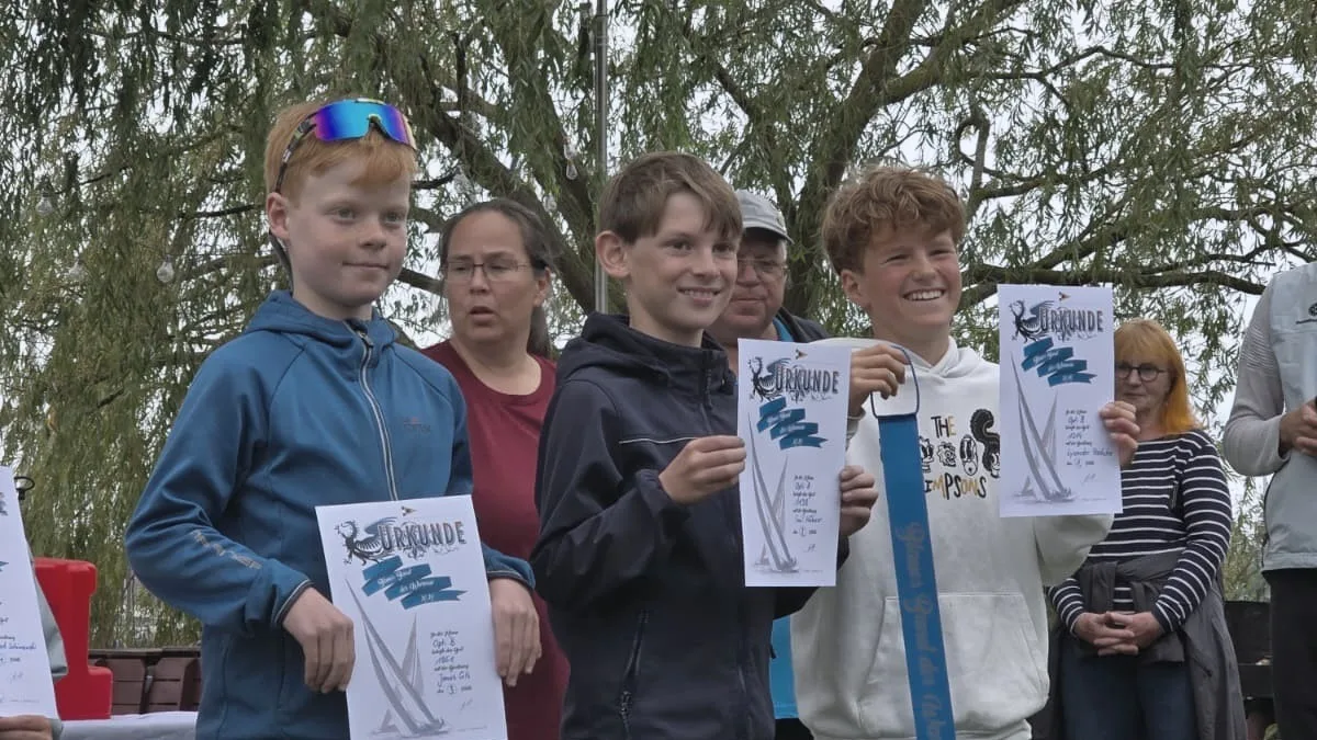Tres regatistas jóvenes con diplomas en un pantalán, uno con la cinta azul de la regata al cuello, un sauce al fondo