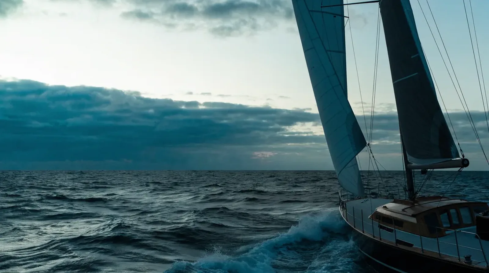 Velero navegando a vela en mar abierto y movido al atardecer, mayor y vela de proa frente a un cielo nublado, espuma rompiendo en la proa.