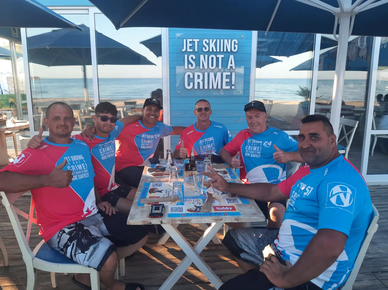 Seis pilotos de motos de agua con camisetas de equipo y el logo de NAMMERT en un chiringuito, pulgar arriba