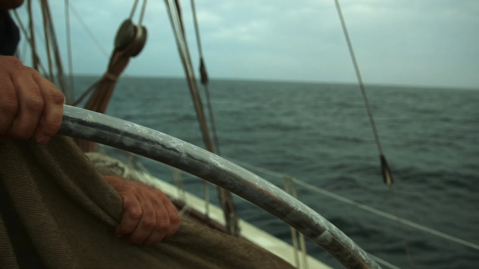 Una mano en la rueda del timón de un velero de crucero, mar abierto al frente bajo cielo cubierto