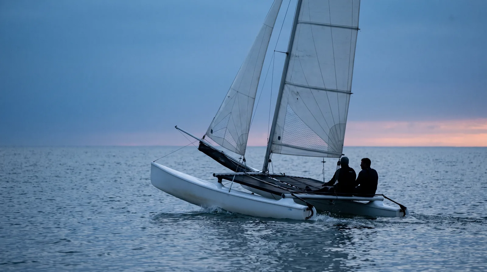 Catamarán de playa navegando al anochecer, con dos tripulantes sobre la red