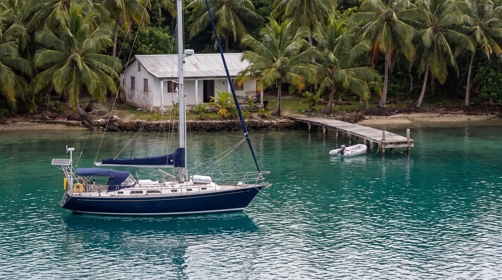 Velero de crucero fondeado en una bahía tropical con una pequeña clínica en la orilla