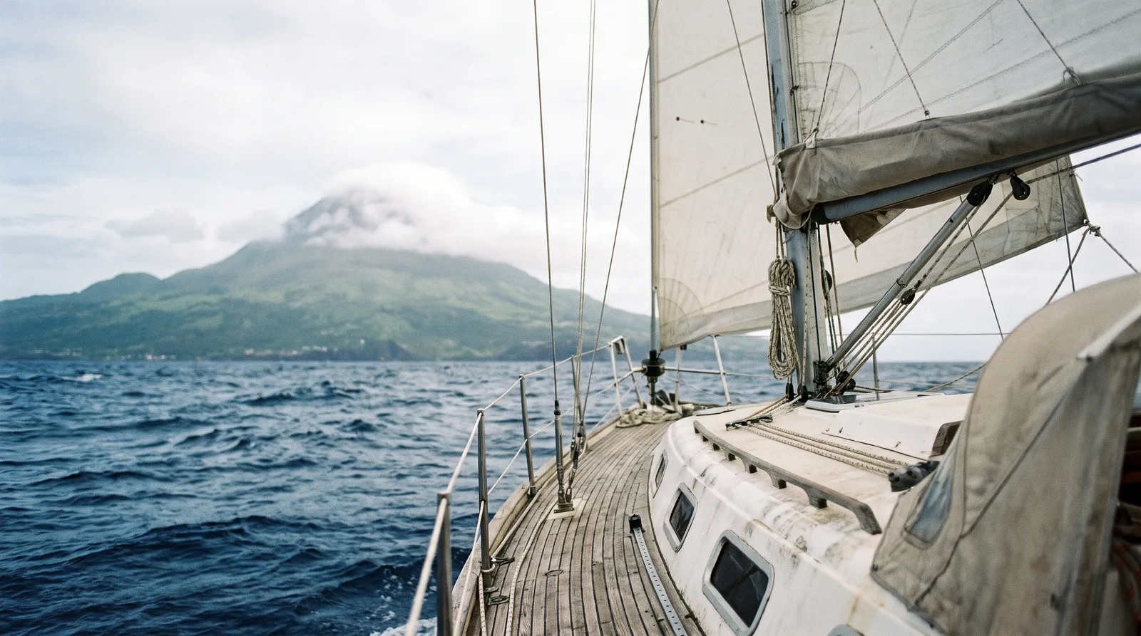 Vista desde la proa de un velero con la mayor rizada sobre el mar de fondo del Atlántico hacia una isla volcánica verde con la cumbre entre nubes