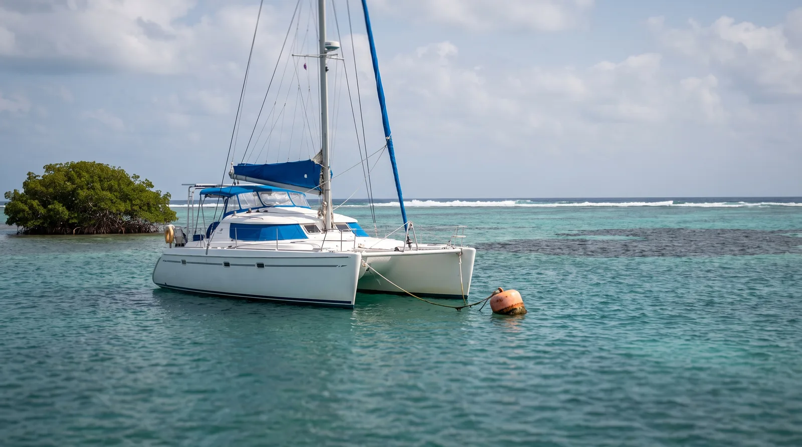 Catamarán de vela cuidado amarrado a una boya en una laguna dentro del arrecife, junto a un cayo de manglar y con rompientes en el horizonte