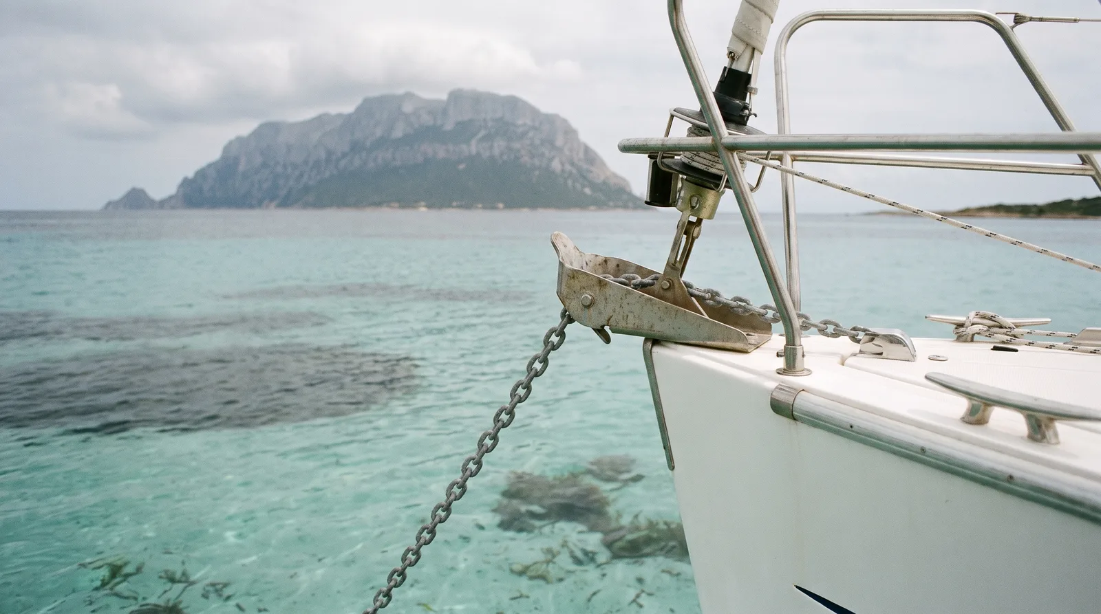 Proa de un velero con la cadena del ancla sobre arena clara y la isla de Tavolara al fondo