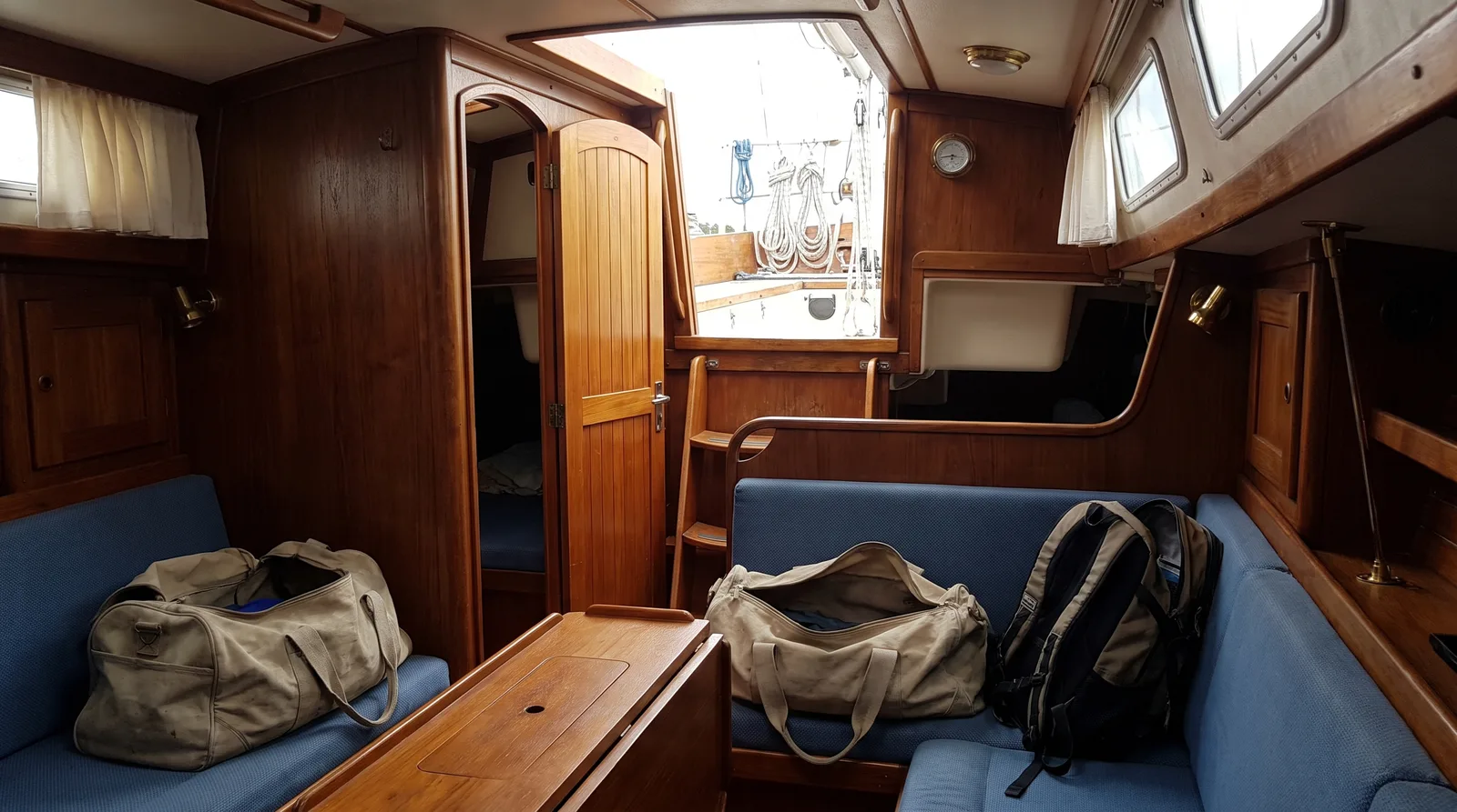 Salón de un velero con carpintería de madera, bolsas de viaje abiertas y una mochila sobre los sofás, la entrada abierta