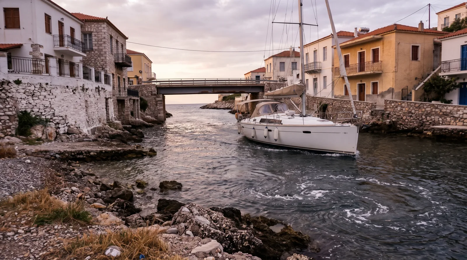 Velero a motor en un canal estrecho entre casas griegas con balcones, detrás un puente bajo y remolinos de corriente en el agua