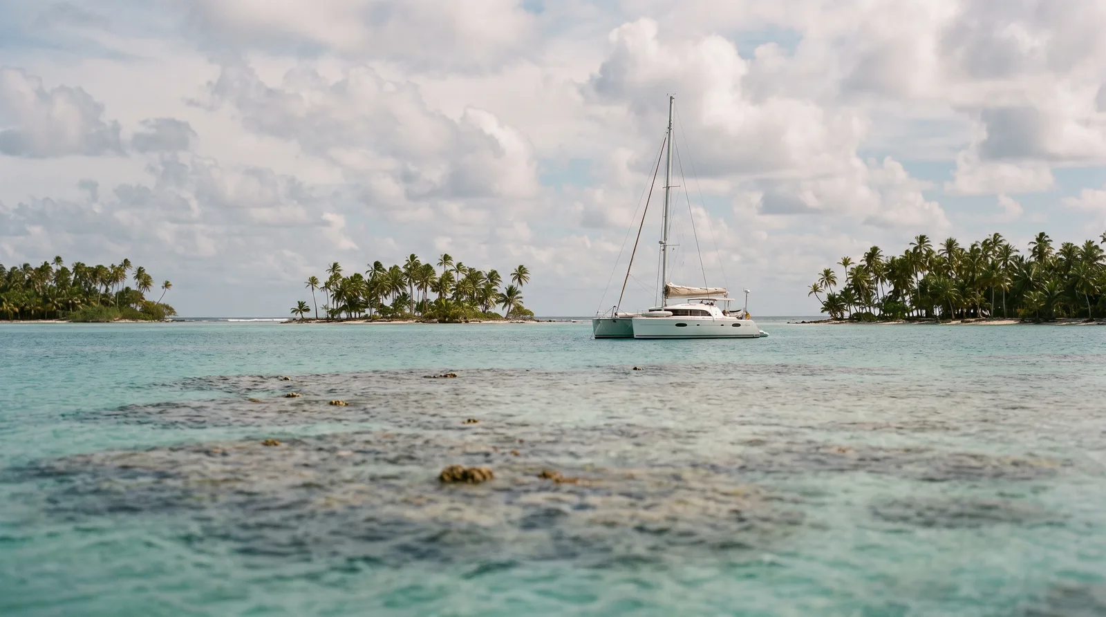 Catamarán de vela en una laguna turquesa tras un arrecife entre islotes con palmeras