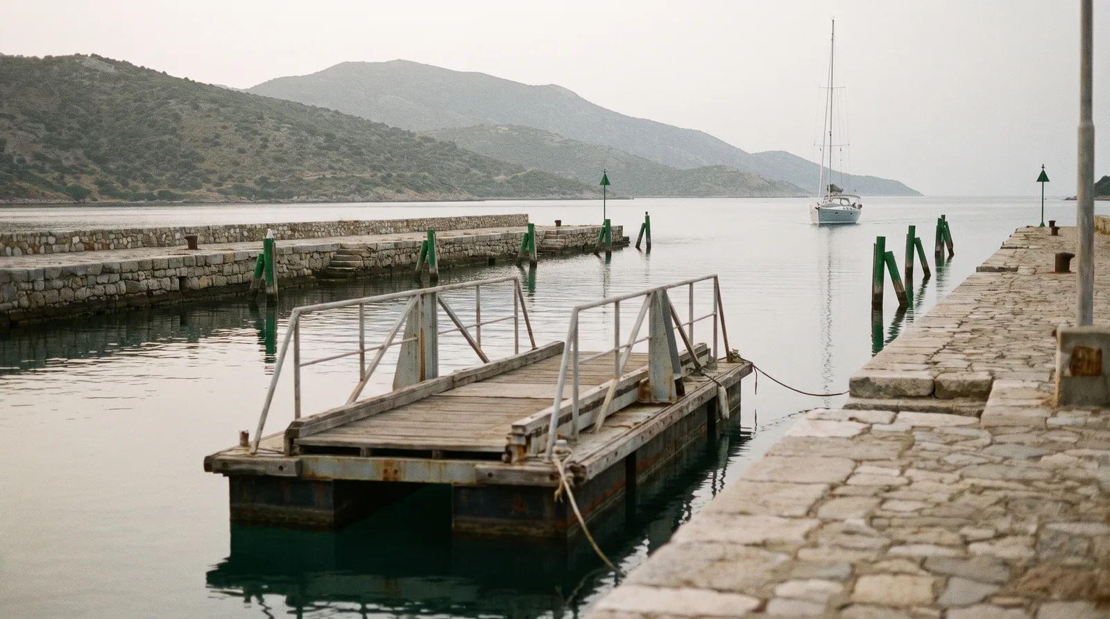 Canal estrecho con orillas de piedra y balizas, un tramo de puente flotante abierto a un lado y un velero bien cuidado a lo lejos