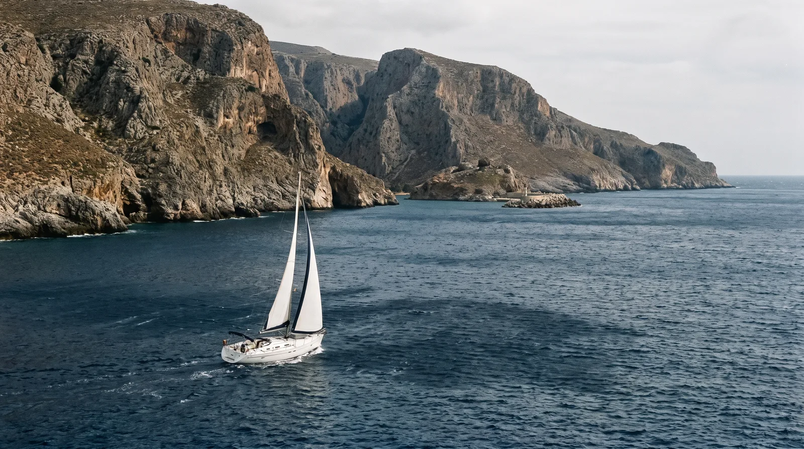 Velero blanco navegando bajo la costa rocosa y escarpada del sur de Creta con barrancos, manchas oscuras de rachas en el agua