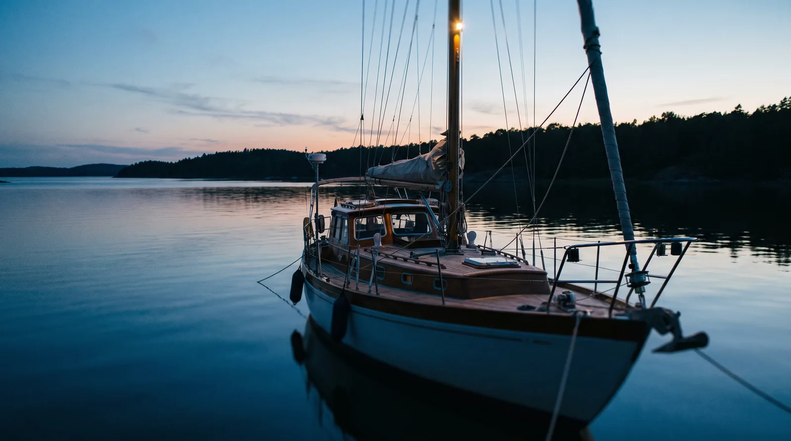 Velero fondeado en una cala resguardada a la hora azul con la luz de fondeo encendida en el tope del palo