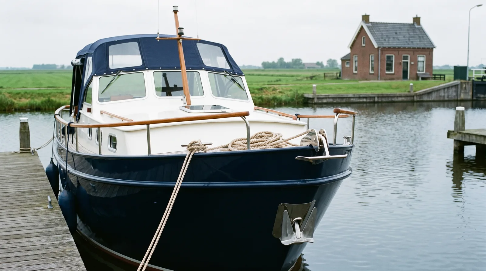 Crucero a motor de acero bien cuidado con capota azul en un pequeño puerto de Frisia junto al IJsselmeer, con pólder llano y una casa de esclusa de ladrillo al fondo