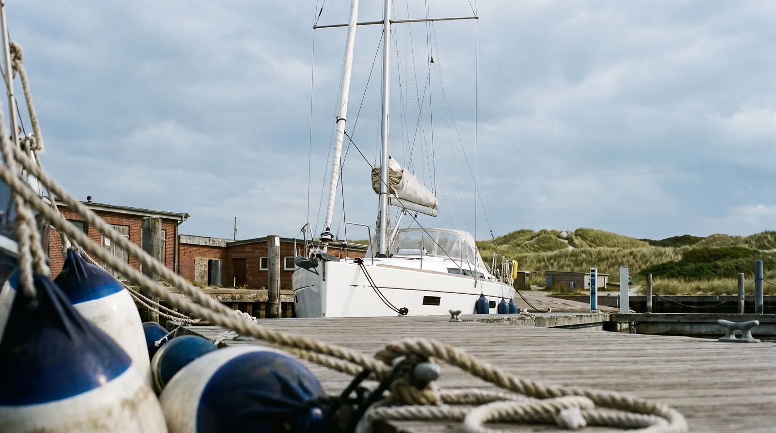 Velero moderno en un pantalán de madera de un puerto del Báltico, con defensas y cabos delante y casas de ladrillo y dunas al fondo