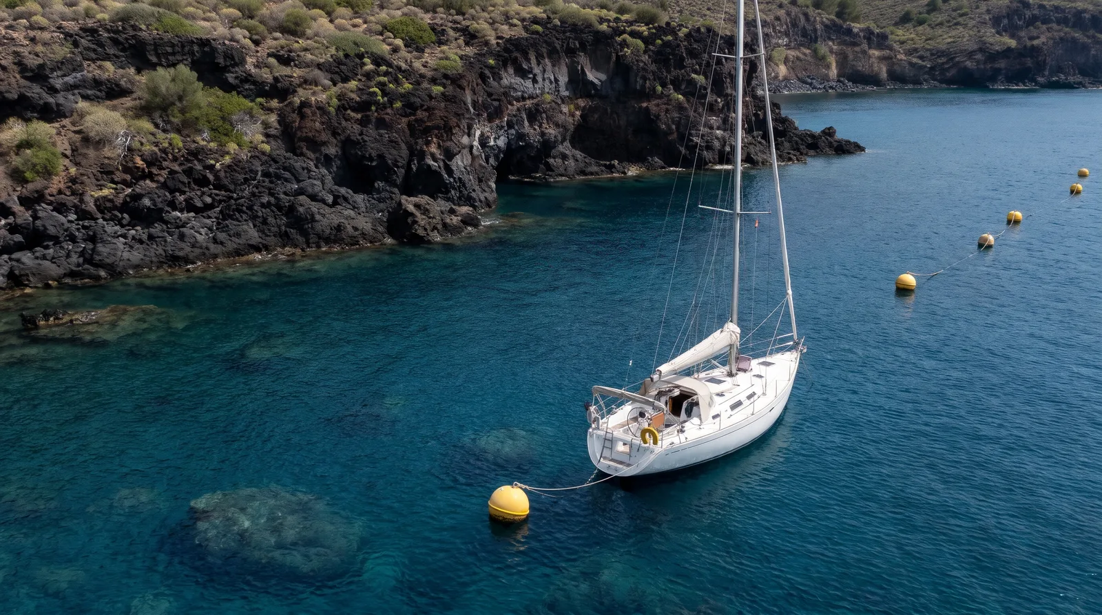 Velero blanco amarrado a una boya amarilla frente a una costa de lava negra, en agua azul intenso y transparente