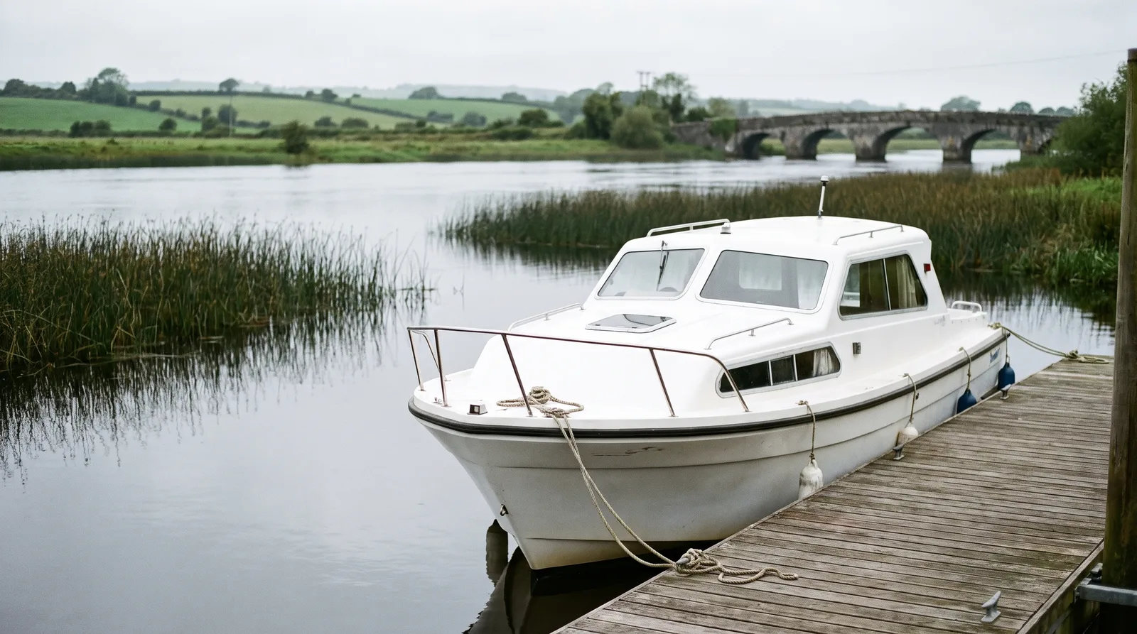 Lancha blanca de alquiler amarrada a un pantalán de madera en un río ancho y tranquilo de Irlanda con juncos, prados y un puente de piedra