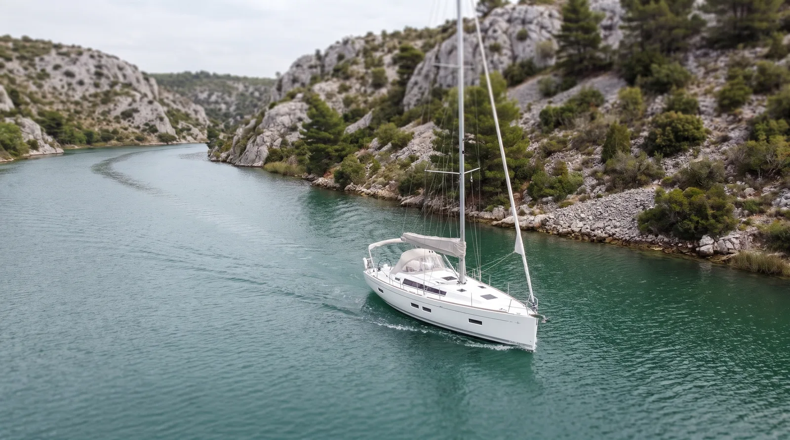 Velero blanco a motor en un río verde y tranquilo entre laderas de caliza clara con pinos y matorral