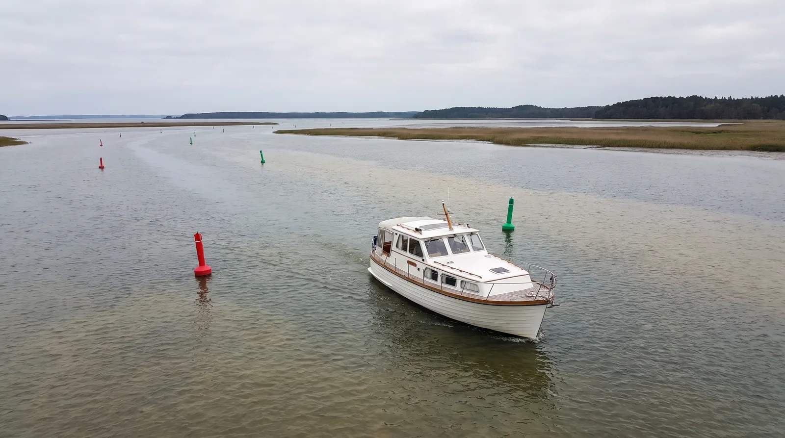 Barco a motor blanco avanzando despacio entre boyas rojas y verdes por una laguna amplia y somera con cañaverales y una orilla boscosa baja