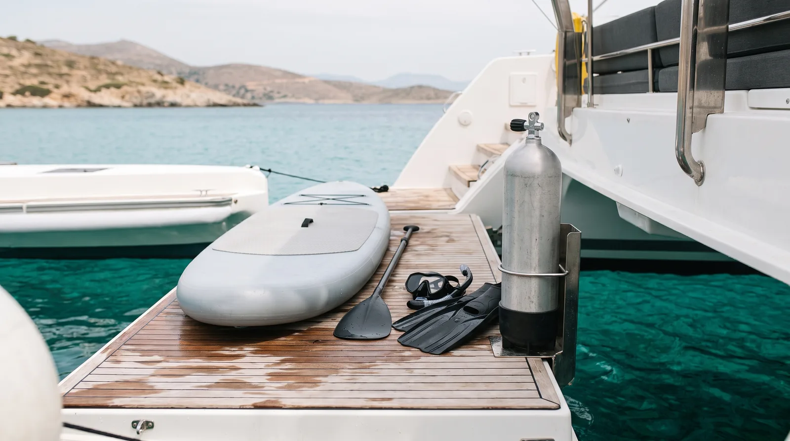 Tabla de paddle surf, pala, gafas, tubo y aletas en la plataforma de baño de un catamarán bien cuidado en una cala de agua clara