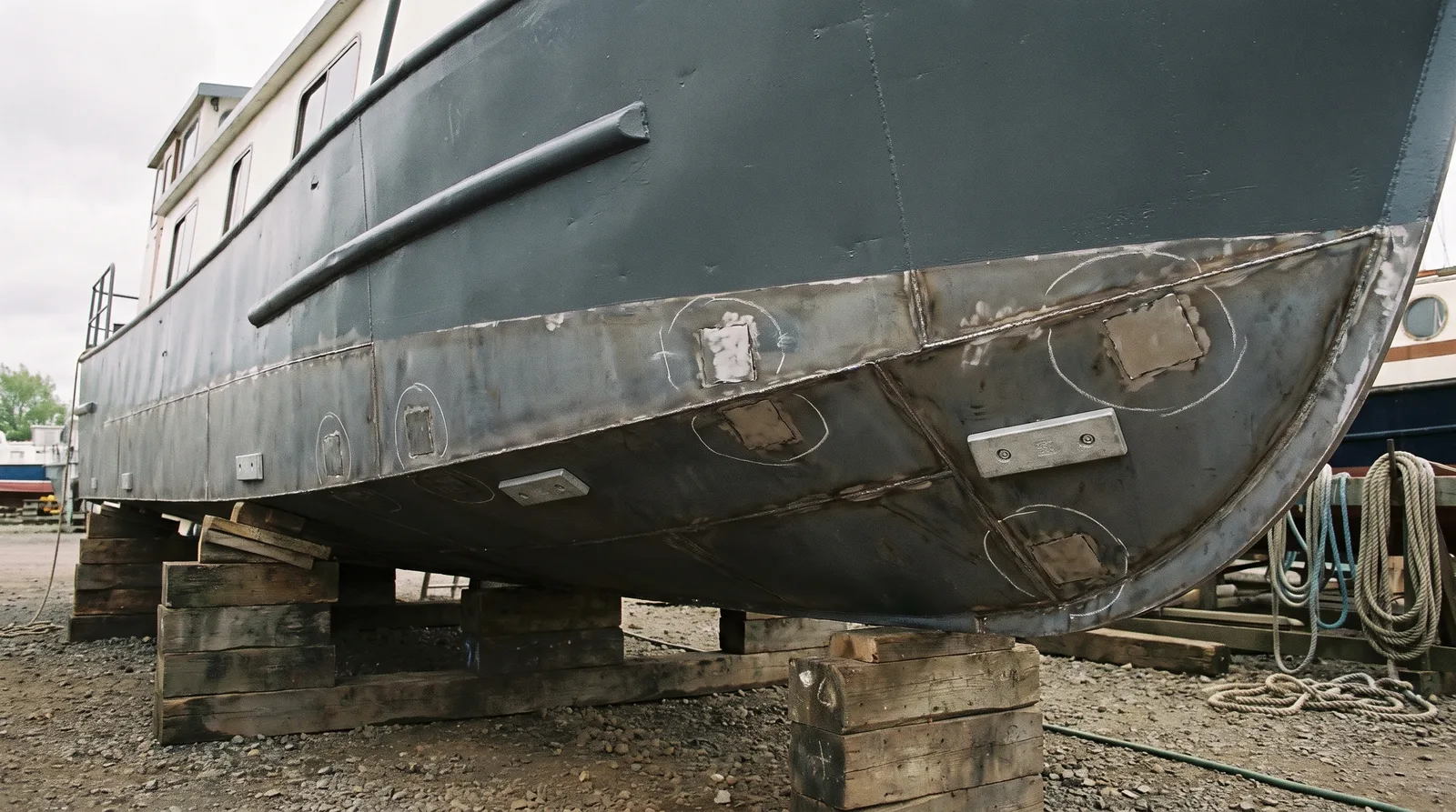 Casco de acero de una casa flotante en seco sobre picaderos de madera, con cordones de soldadura, puntos de medición marcados con tiza y ánodos de sacrificio