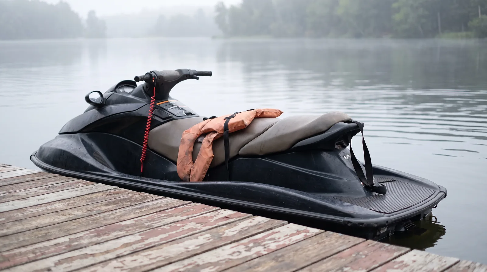 Una moto de agua vacía con un chaleco sobre el asiento y el cordón rojo de hombre al agua en el manillar, junto a un pantalán de madera con bruma