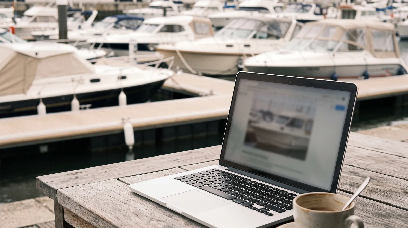 Portátil con un portal de compraventa de barcos sobre una mesa en el puerto, al fondo lanchas bien cuidadas en el pantalán