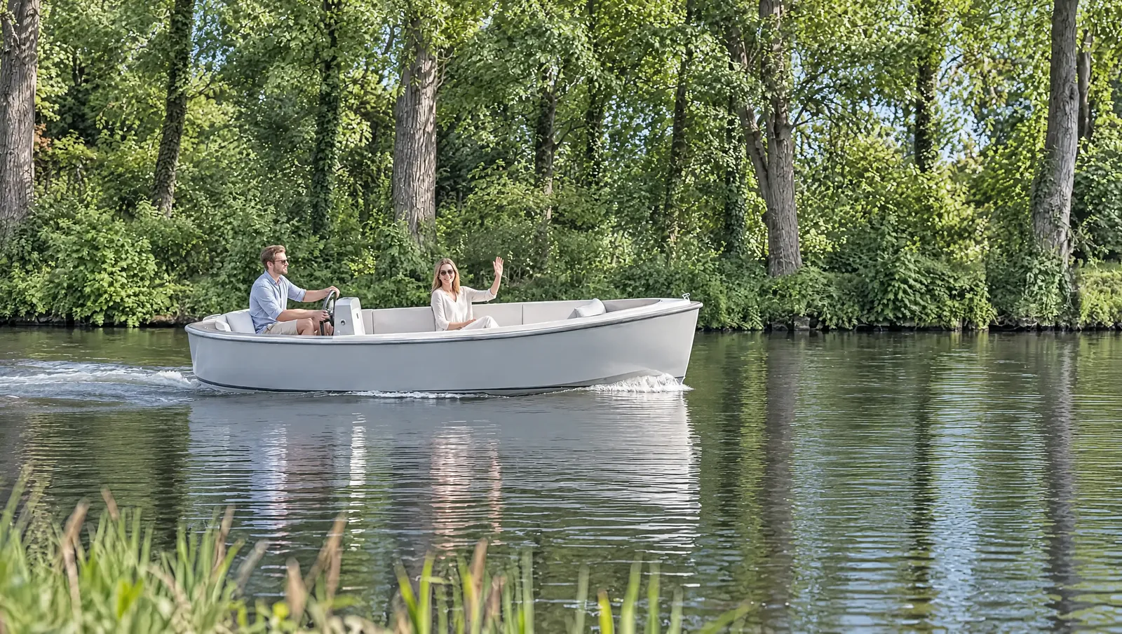 Barco eléctrico gris claro navega por un canal arbolado a pleno sol, él lleva el timón y ella ríe y saluda a la orilla
