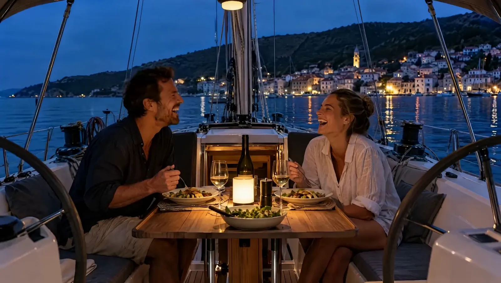 Una pareja ríe durante la cena en la bañera de un velero, las luces del pueblo brillan sobre el agua